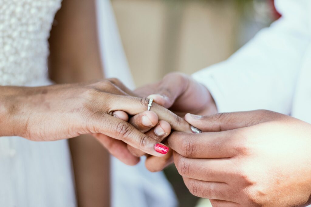 The picture is a close up of a couple's hands. The person on the right is placing an engagement ring on the finger of the person on the left. Her fingernails are painted a sparkly red and silver. They are both wearing white blurred in the background.