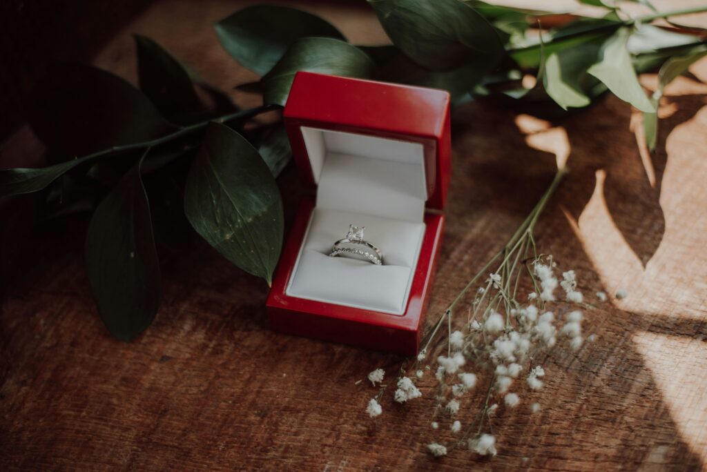 An engagement ring sits on a white cushion inside a red box. It's on a shiny, dark wood surface in between baby's breath flowers and generic dark green flower stems and leaves.