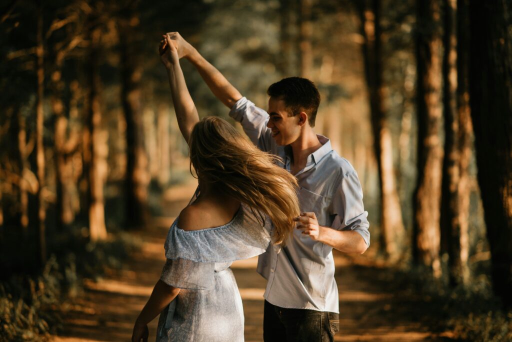 A couple dances in the woods. The trees cast large shadows, indicated the day is close to dusk. Both are wearing pastel blue tops, and the man twirls the woman under his arm with a smile.